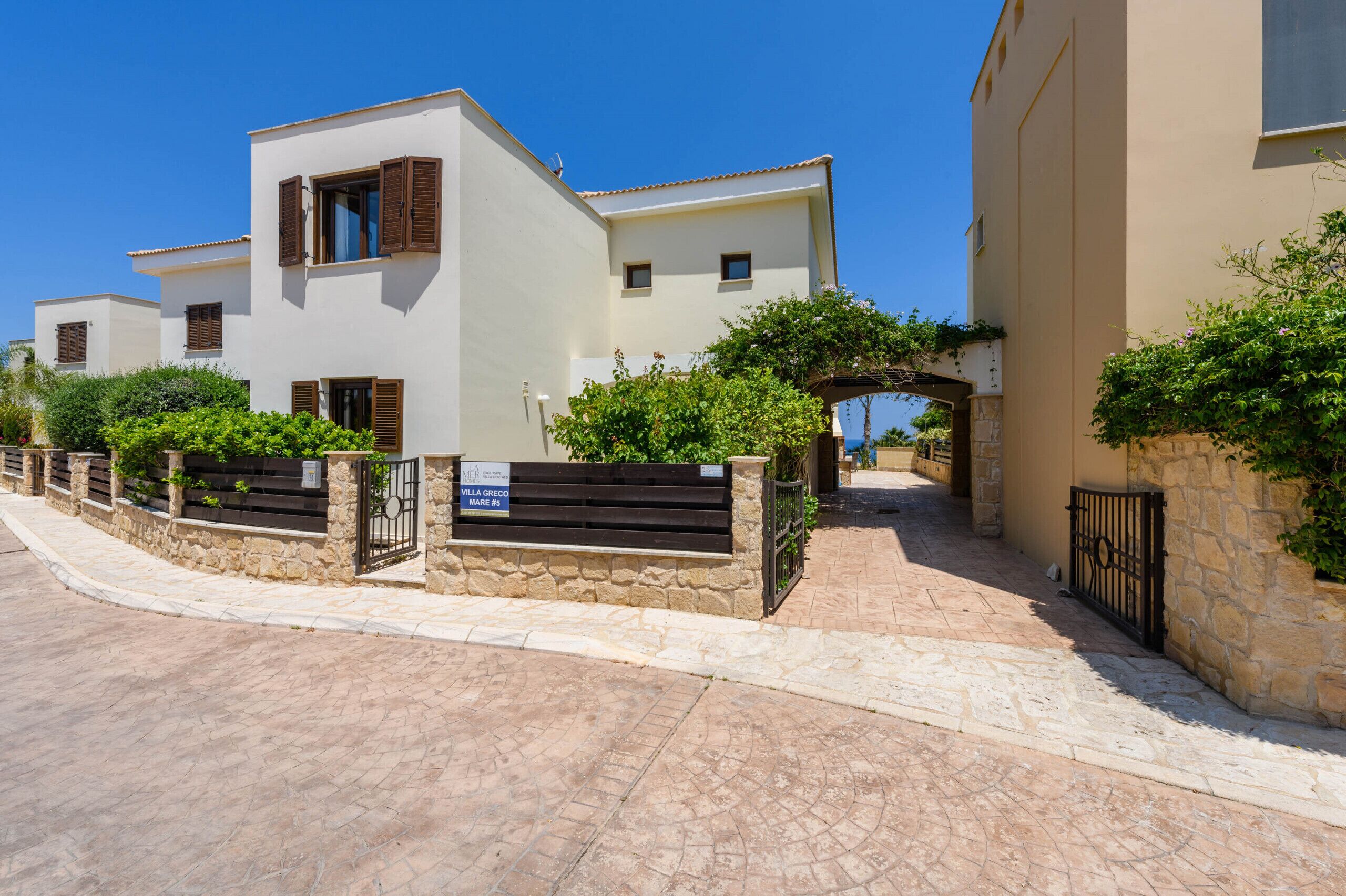 Calle residencial con edificios blancos, muros de piedra y un cartel que indica 'Villa Greco Monte 24'.