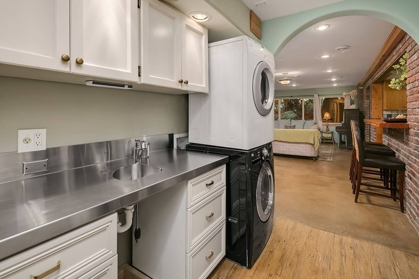 A compact kitchen with stainless steel countertops, a built-in washing machine, and a dining area with a brick wall and wooden furniture.