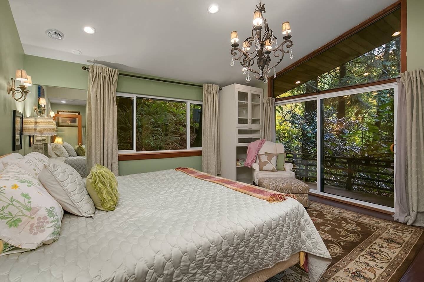 Une chambre avec un grand lit, un lustre, une fenêtre donnant sur un paysage verdoyant et un fauteuil près de la fenêtre.