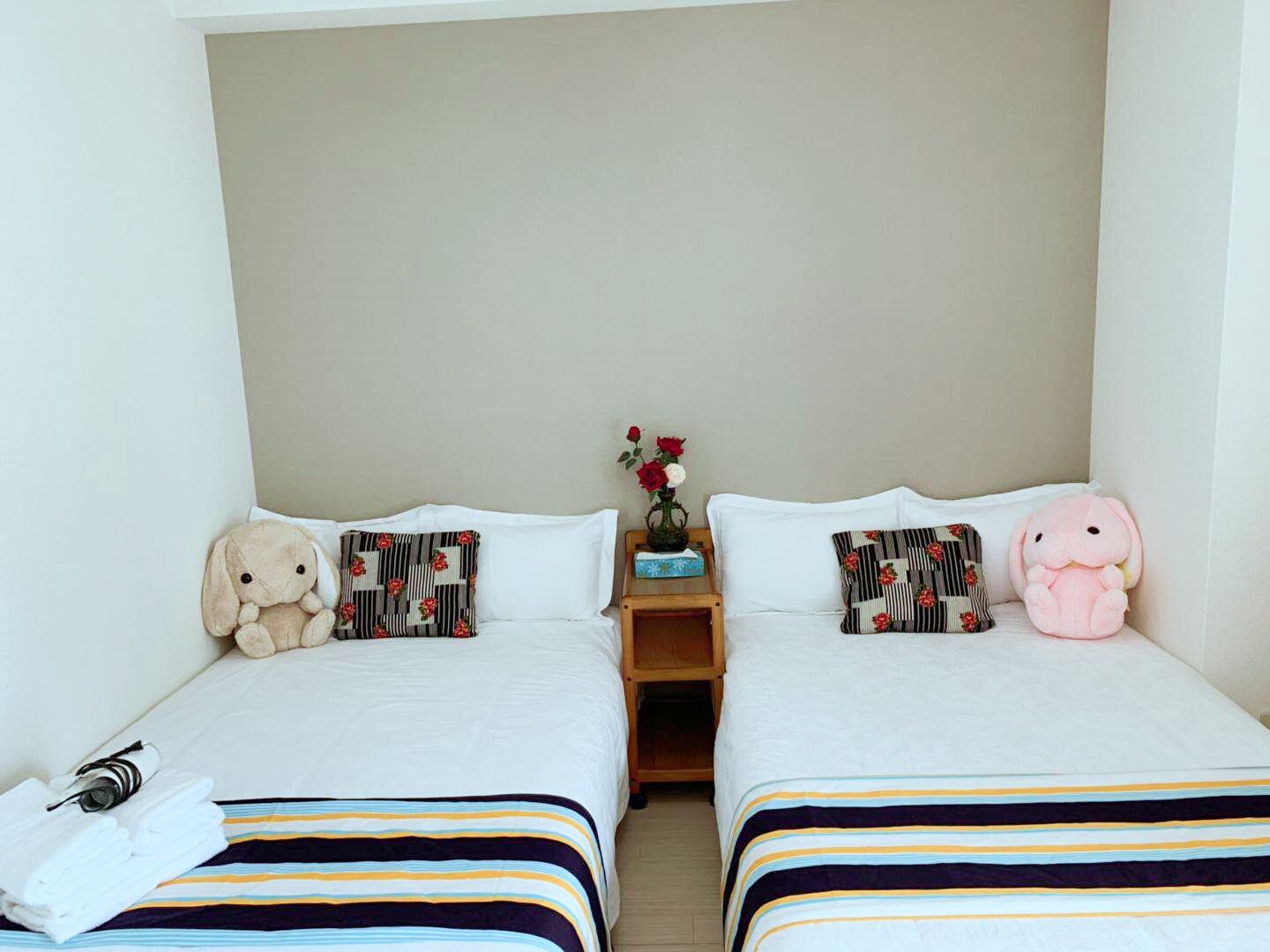 A hotel room with two beds, striped bedding, a nightstand with a vase of flowers, and plush toys on the beds.