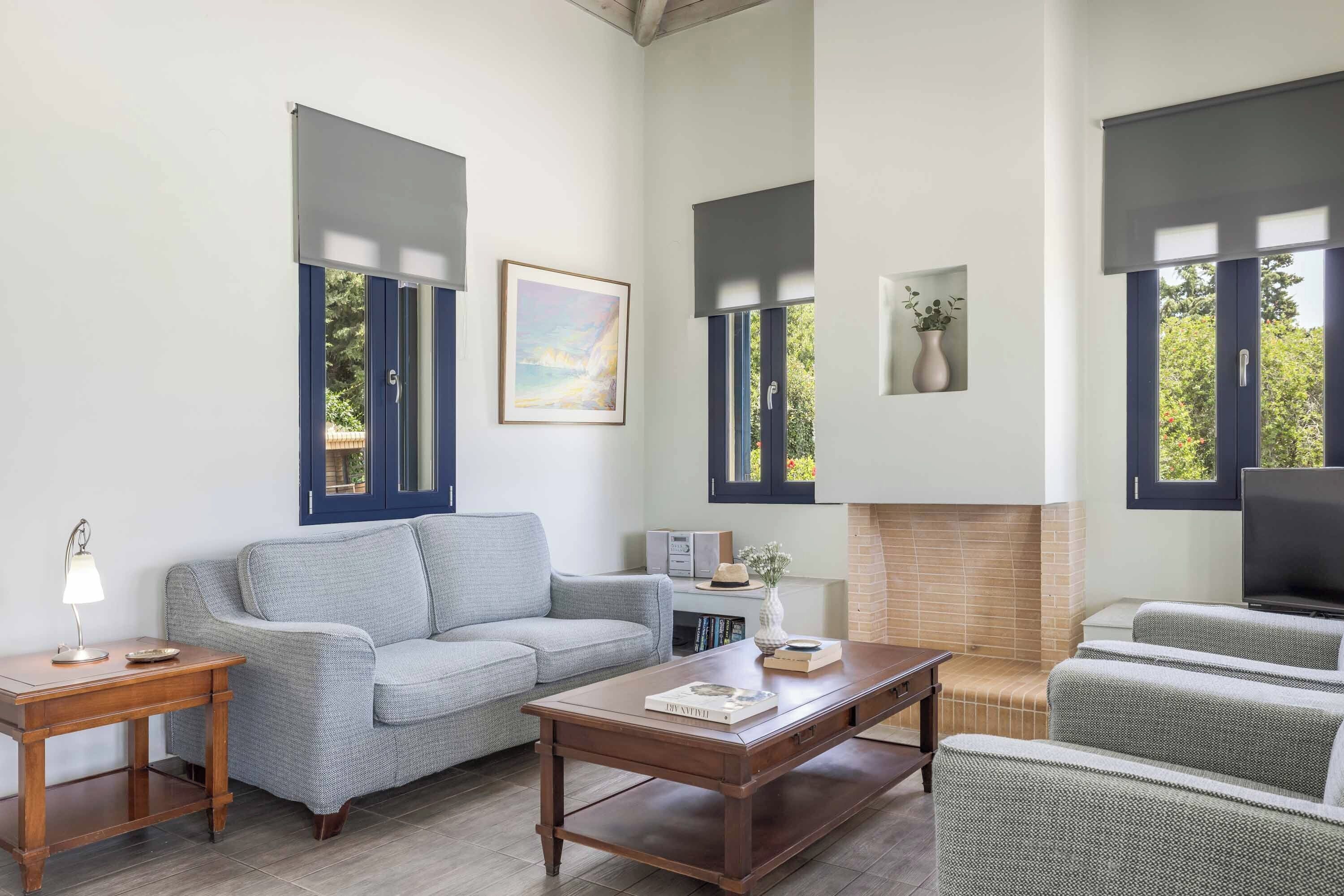 Un salón con sofá, una mesa de centro, chimenea y ventanas grandes con contraventanas.