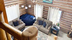 Interior acogedor de una cabaña con pared de troncos de madera, un sillón de peluche azul, dos sofás con cojines, una mesita con una planta y un cuadro de un barco en la pared.