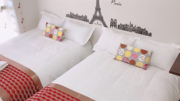 Habitación de hotel con dos camas, cada una con ropa blanca y almohadas coloridas. Un edredón blanco y rojo a rayas se ve en el suelo. La pared tiene un mural con un paisaje urbano que incluye la Torre Eiffel y la palabra 'Paris'.