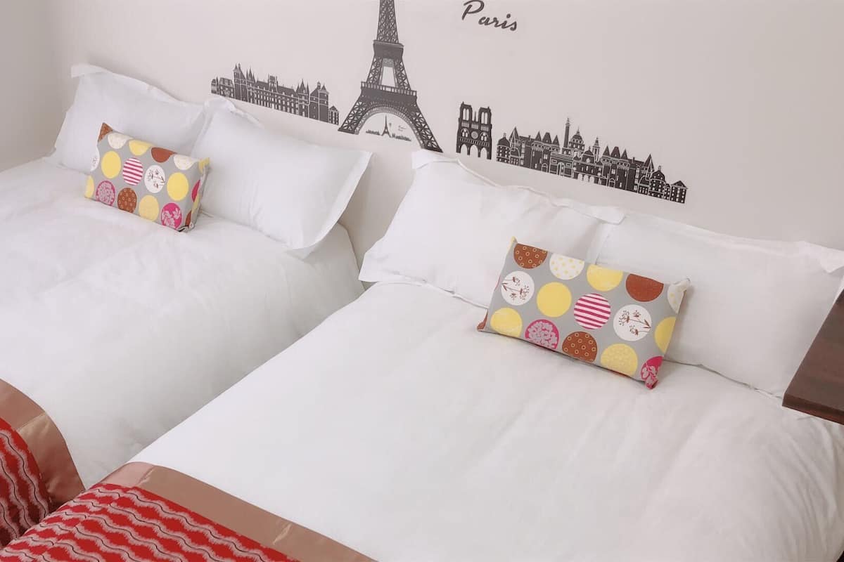 Habitación de hotel con dos camas, cada una con ropa blanca y almohadas coloridas. Un edredón blanco y rojo a rayas se ve en el suelo. La pared tiene un mural con un paisaje urbano que incluye la Torre Eiffel y la palabra 'Paris'.