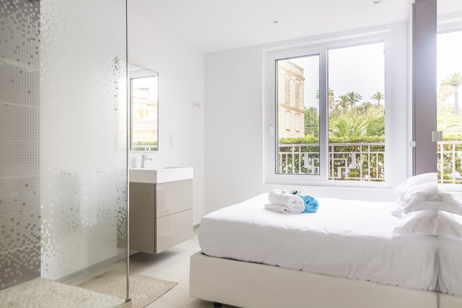 A modern hotel room with a large bed, a bathroom with a glass shower, and a balcony with a view of palm trees and buildings.