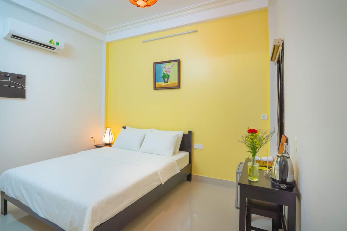 A neatly made bed in a room with a yellow wall, a small table with a vase of flowers, and an air conditioning unit on the wall.