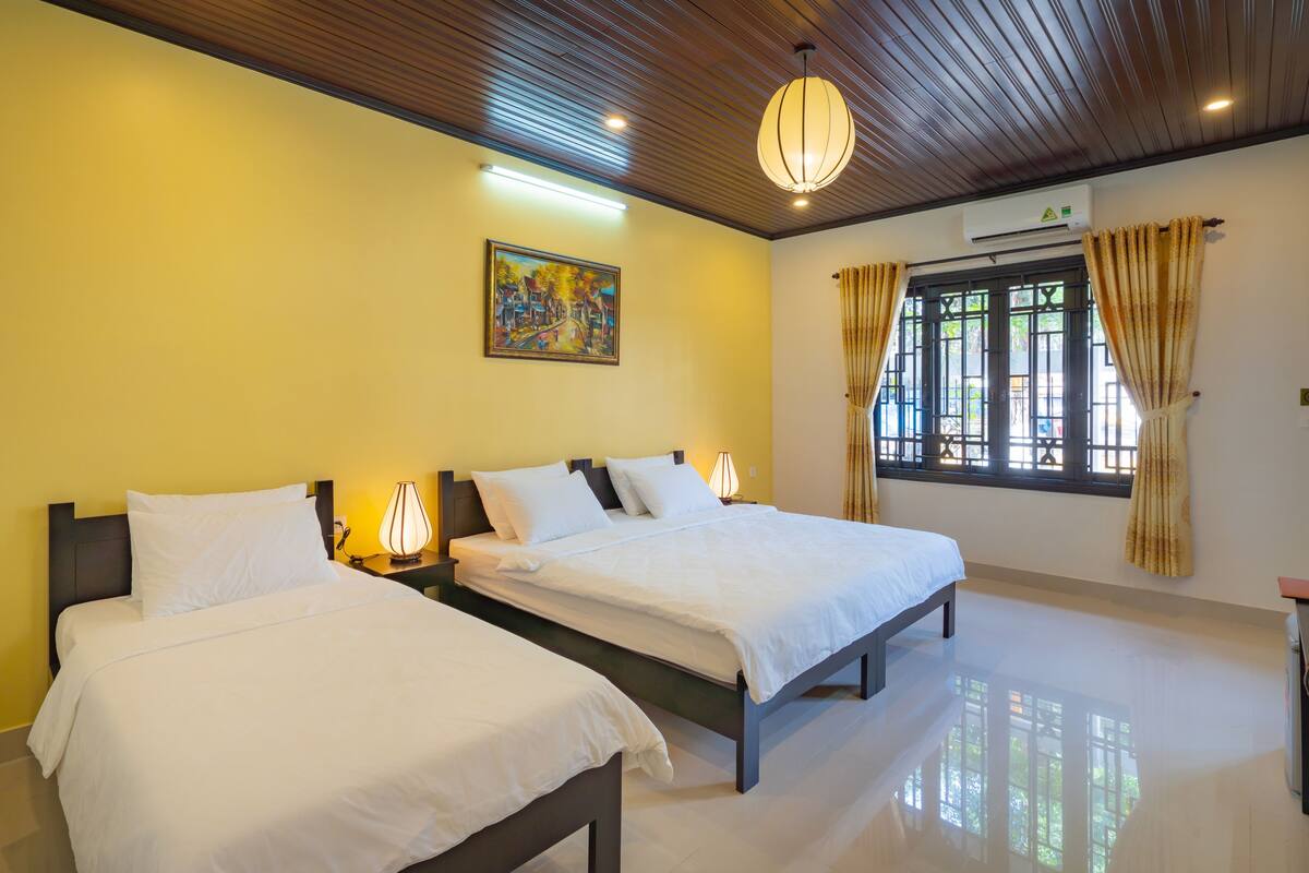 A hotel room with two beds, a painting on the wall, a window with curtains, and an air conditioning unit.