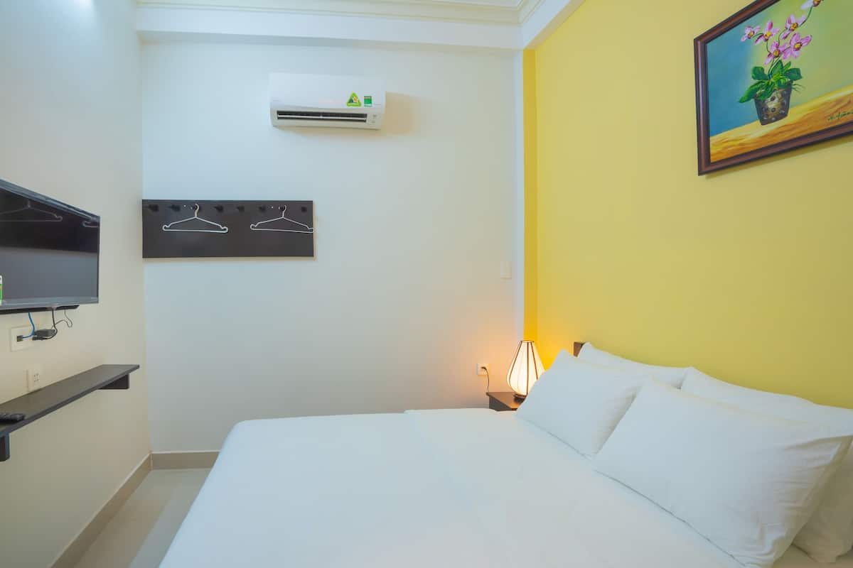 A hotel room with a bed, a TV mounted on the wall, an air conditioning unit, a wall-mounted coat rack, and a painting of flowers.