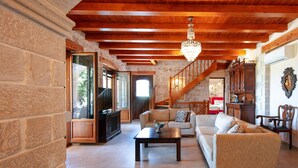 A spacious living room with a stone wall, wooden ceiling beams, a chandelier, a sofa, a coffee table, and a staircase leading to another level. - Villa Melpomeni | in a Forest with Private Pool, Sea View, Gym & Home Cinema (Rethymno)