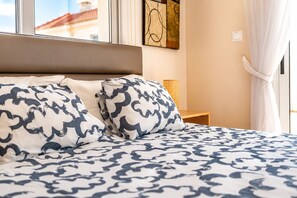 A bed with patterned bedding, a bedside table with a lamp, and a window with curtains. - Narcissos 'Nissi Beach' Apartment D12 (Ayia Napa)