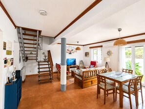 A spacious living area with a wooden staircase, a dining table with chairs, a television, and a large window with a view of greenery.