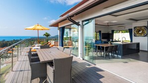 Eine moderne Terrasse im Freien mit Holztisch und Korbstühlen, einem gelben Sonnenschirm und Blick auf den Ozean.