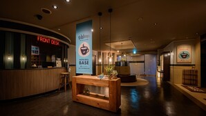 A hotel lobby with a reception desk labeled "FRONT DESK," a central wooden table with decorative items, and a "HOTELWILL BASE" sign. - HOTEL WILL BASE Tsurumi - Adults Only (Yokohama)