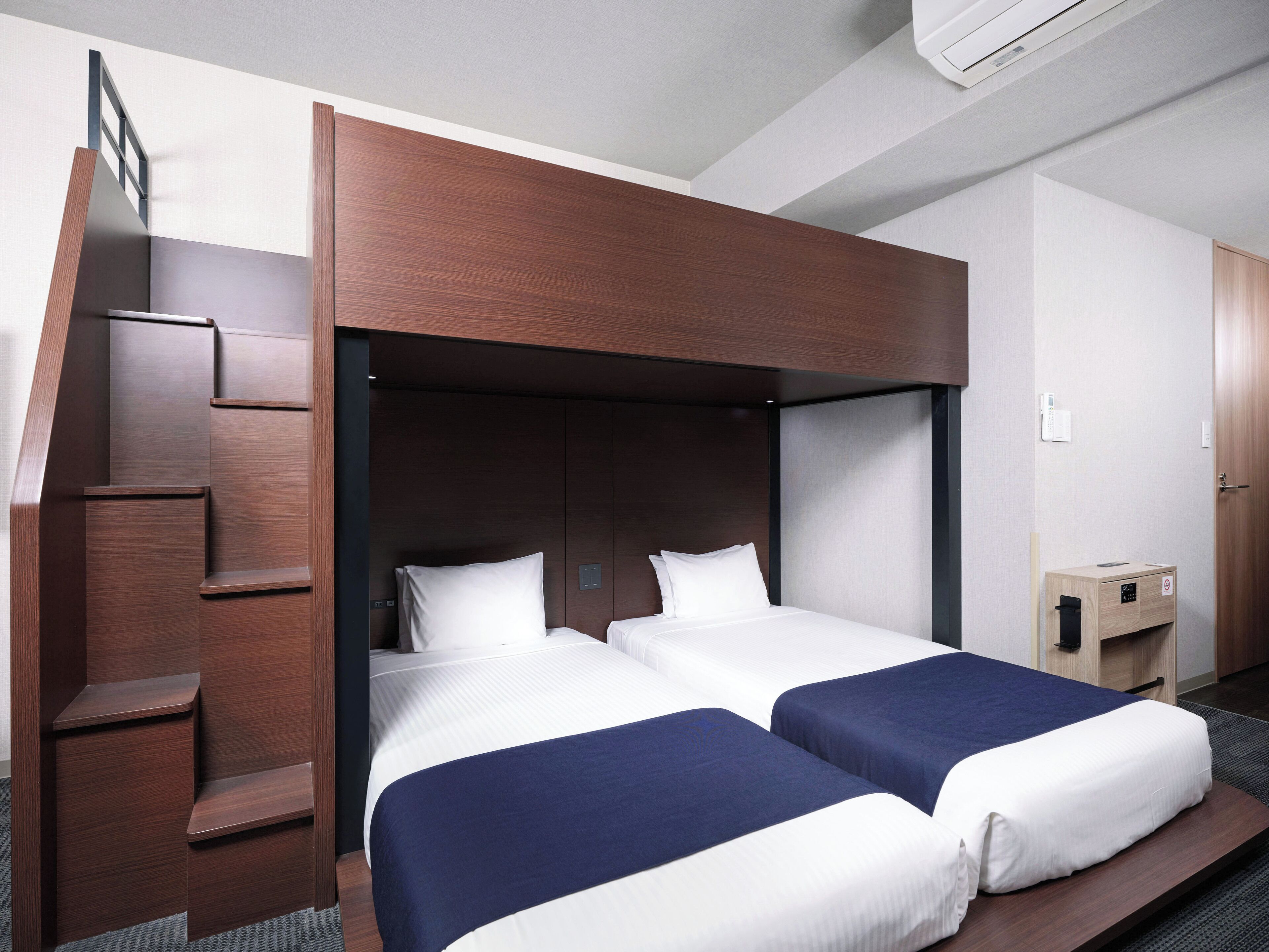 Habitació d'hotel amb dos llits, un panell de capçalera de fusta, una taula de nit de fusta petita i un armari encastrat.