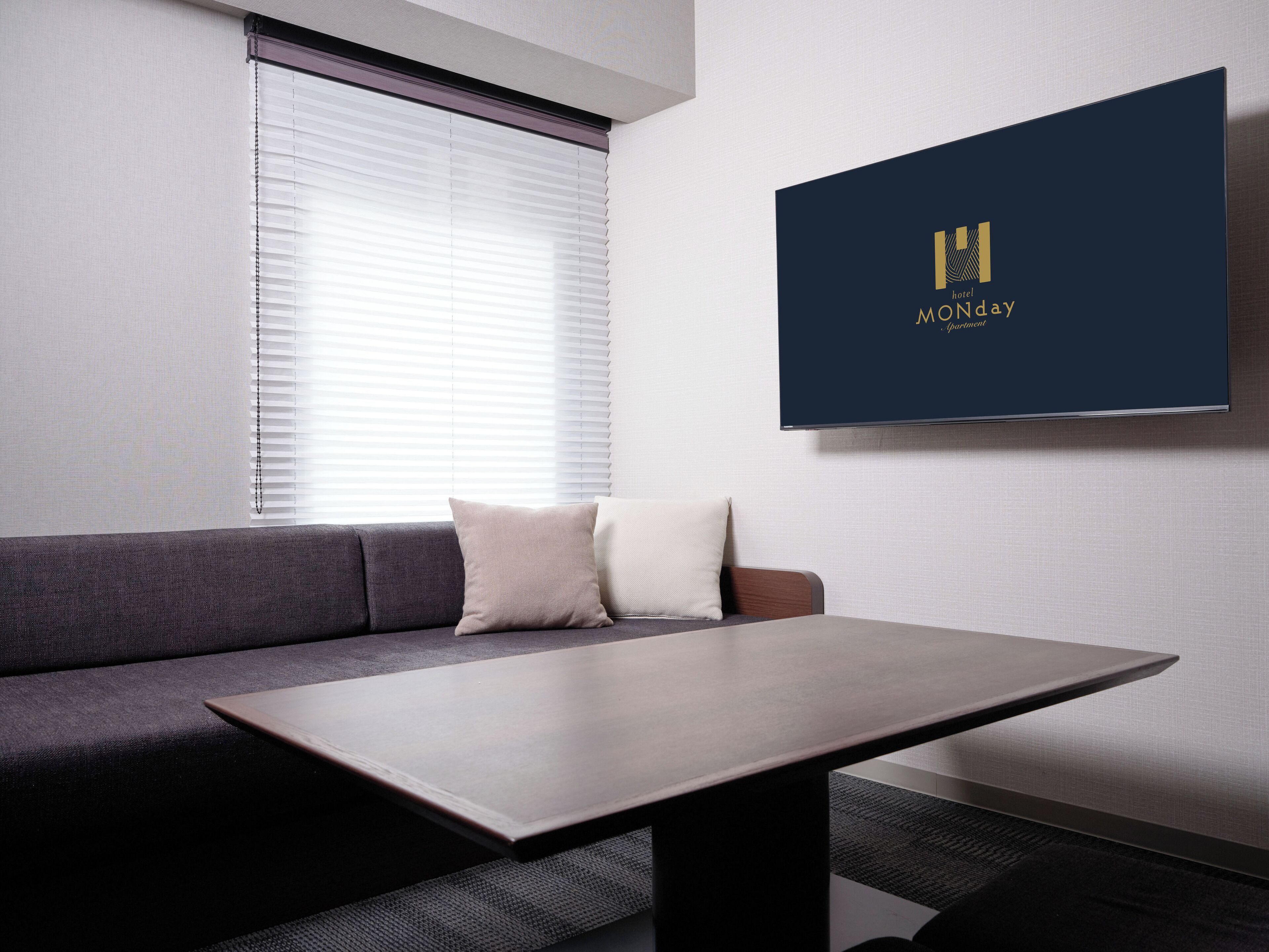 A modern hotel room with a grey sofa, a small table, and a wall-mounted TV displaying the hotel name.