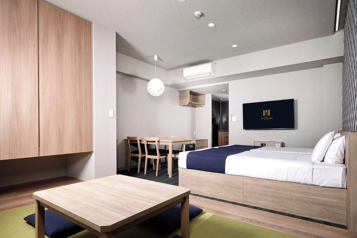 A modern hotel room with a wooden desk, a bed with a blue blanket, a TV on the wall, and a kitchenette in the background.