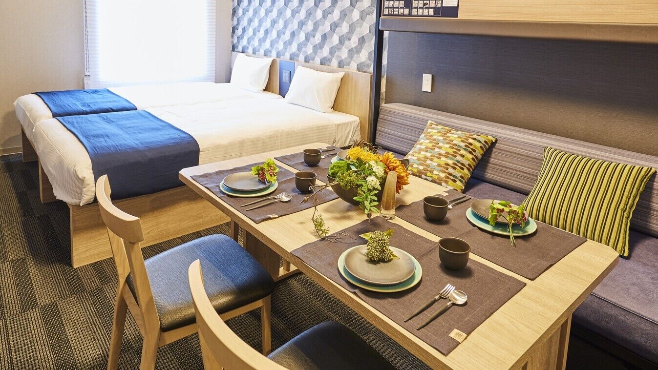 Une chambre d’hôtel avec un lit, une table à manger dressée avec des assiettes, des tasses et des couverts, et un canapé avec des coussins.
