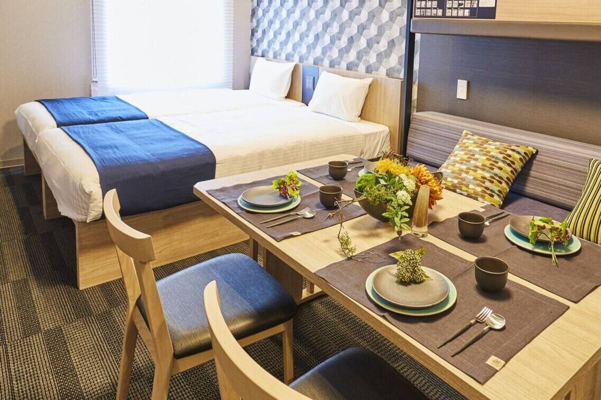 A hotel room with a bed, a dining table set with plates, cups, and cutlery, and a sofa with cushions.