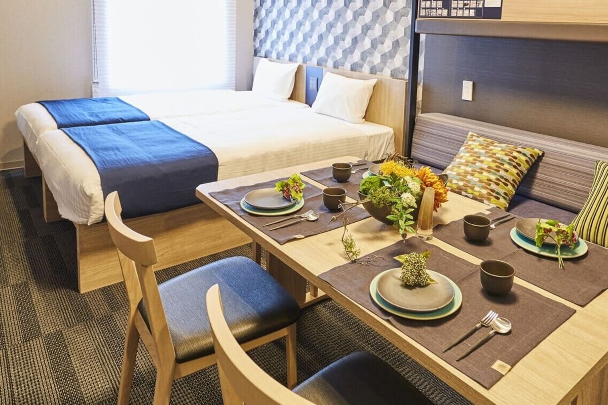A hotel room with a bed, a dining table set with plates, cups, and cutlery, and a sofa with cushions.