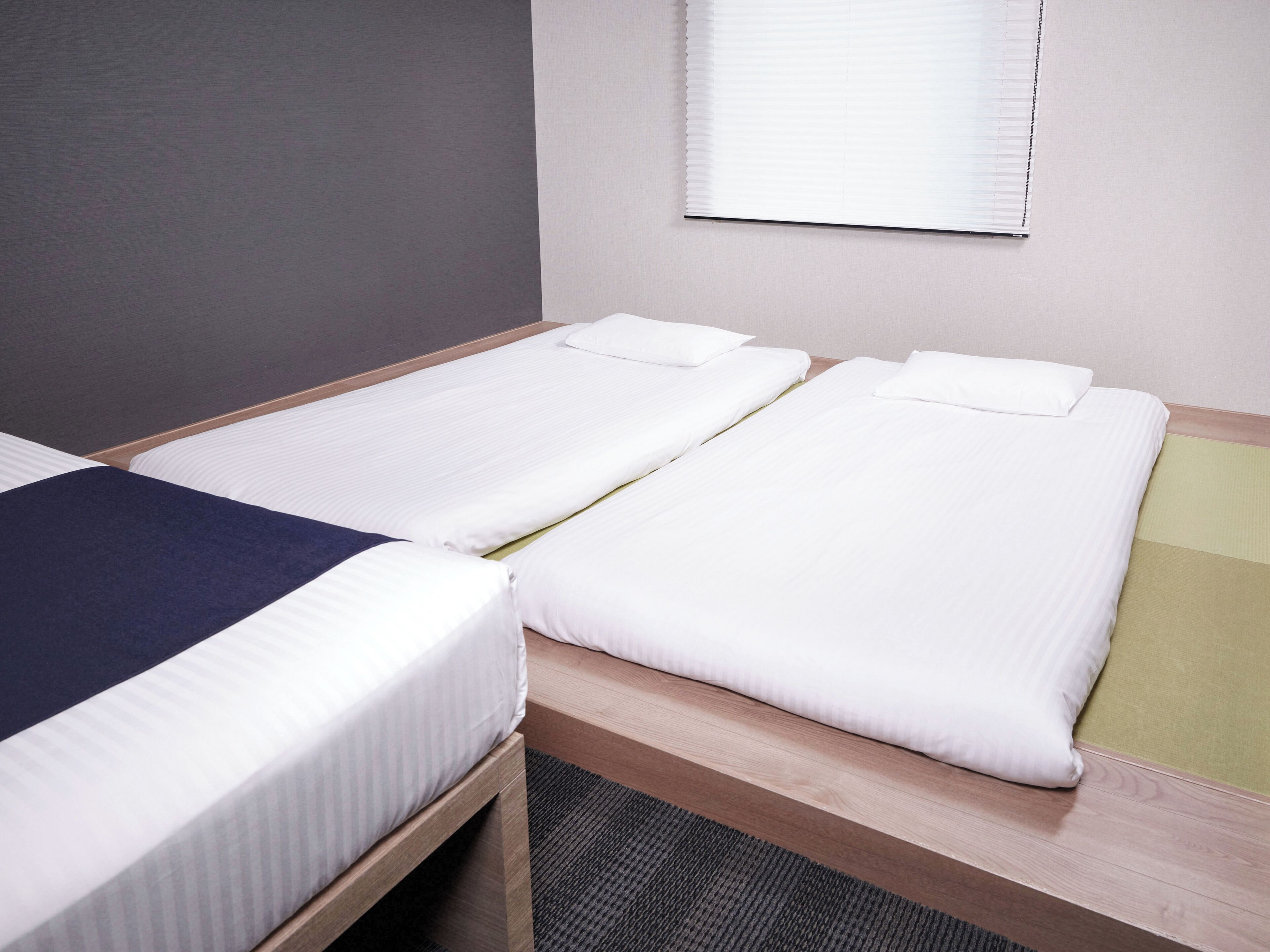 Ein Doppelbett mit blauer Decke und ein Einzelbett mit weißer Decke in einem Zimmer mit Fenster und grauer Wand.