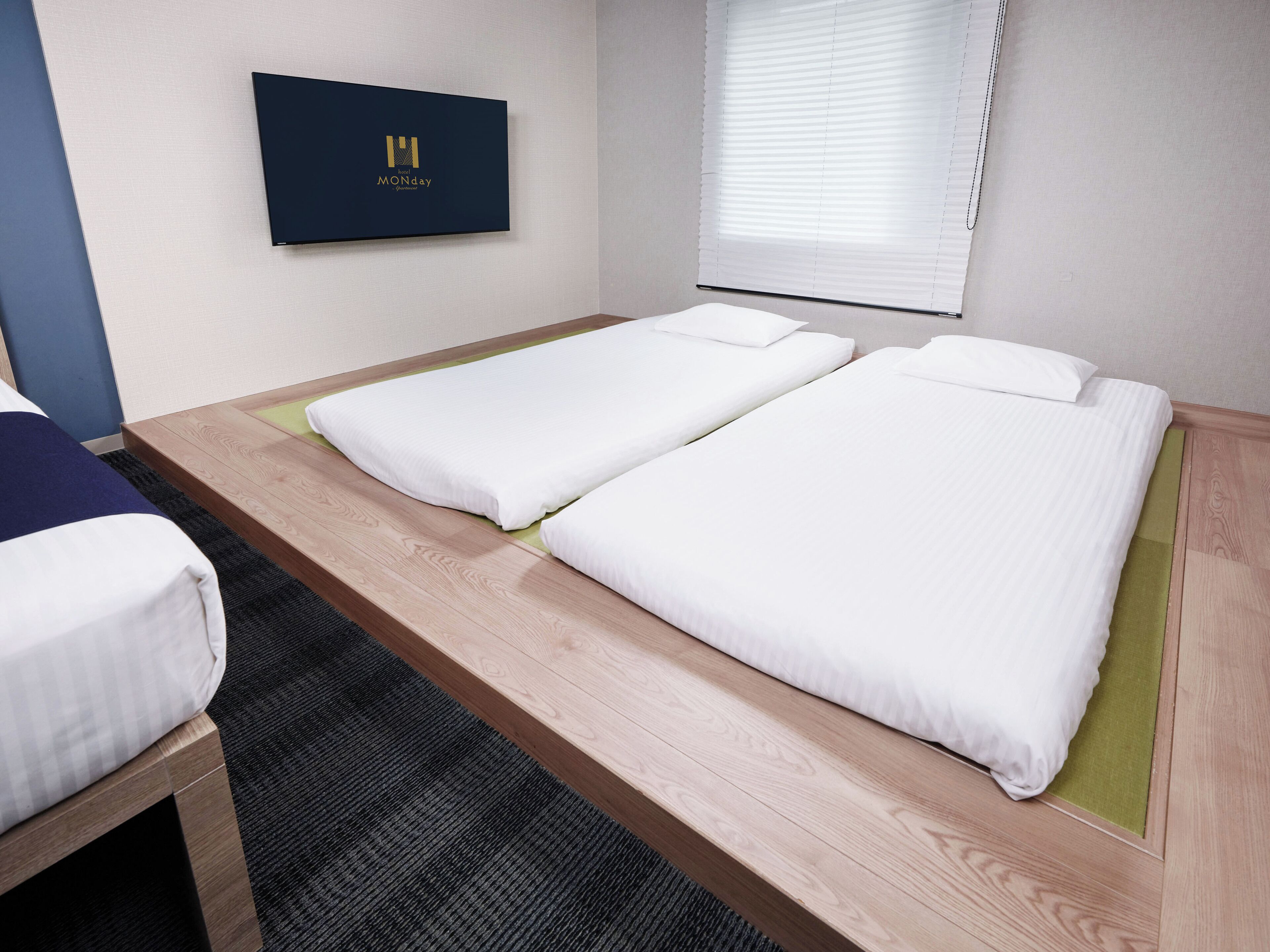 A hotel room with two single beds, a wooden bench, a small table, a framed picture on the wall, and a window with blinds.