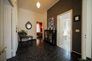 Un pasillo con piso negro moteado, una planta en maceta, una silla negra, una estantería y una puerta que da a otra habitación.