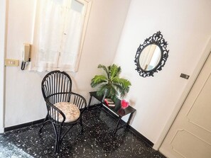 Une pièce avec un sol carrelé de carreaux noirs, une chaise noire avec un coussin à motifs floraux, une petite table sur laquelle est posée une plante en pot, un miroir décoratif et une porte blanche.