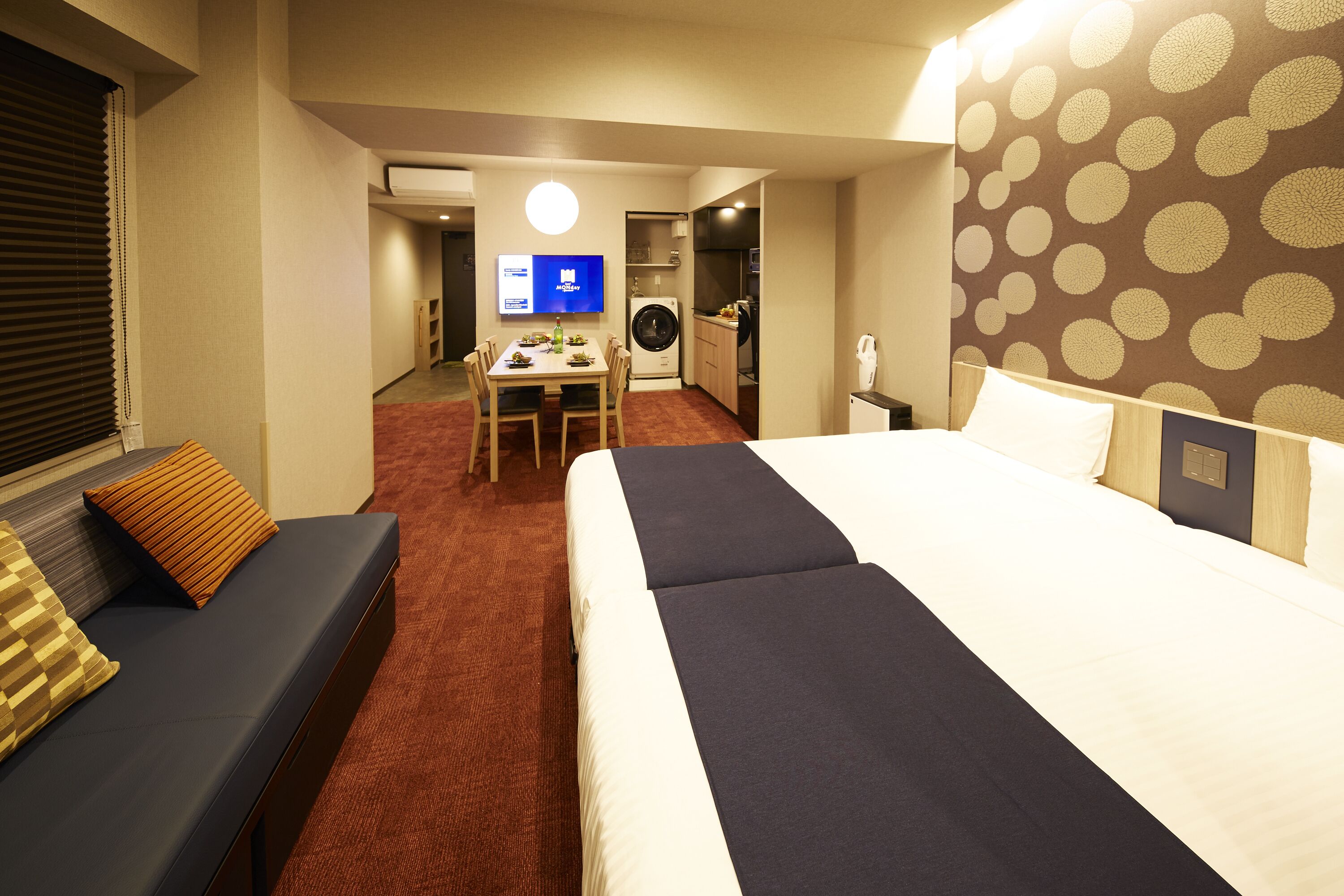 A hotel room with a large bed, a sofa, a dining table with chairs, a washing machine, and a television.