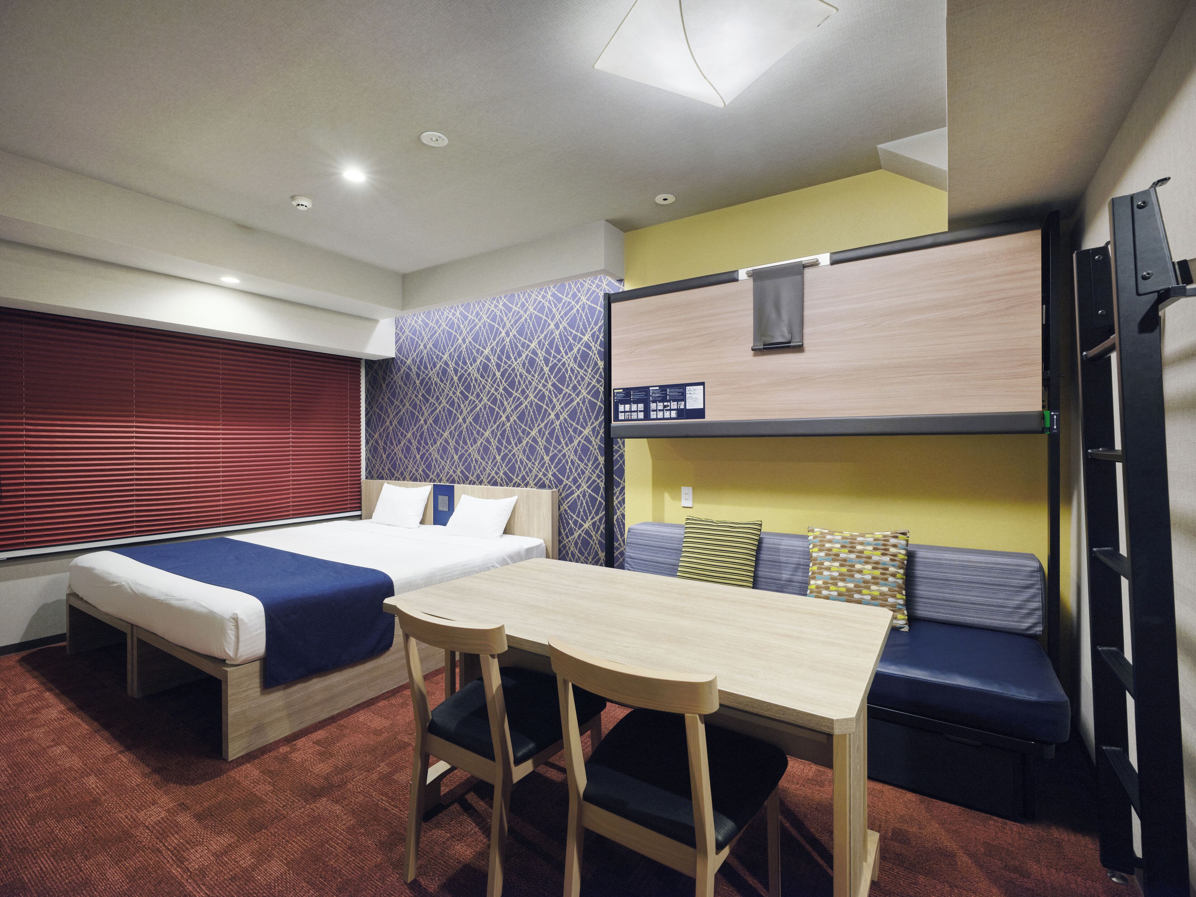 A compact hotel room with a bunk bed, a single bed, a small table with chairs, and a window with blinds.