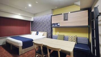 A compact hotel room with a bunk bed, a single bed, a small table with chairs, and a window with blinds.