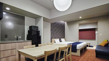A compact hotel room with a kitchenette, a dining area with a table and chairs, a bed with a blue and white bedspread, and a window with red blinds.