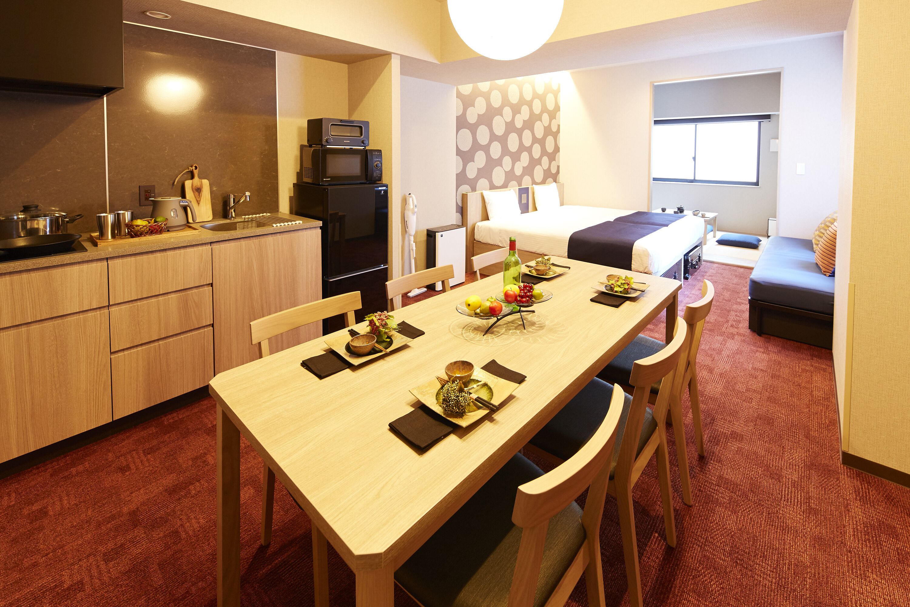 A compact hotel room with a kitchenette, a dining area with a table set for two, a bed with a view, and a small seating area.