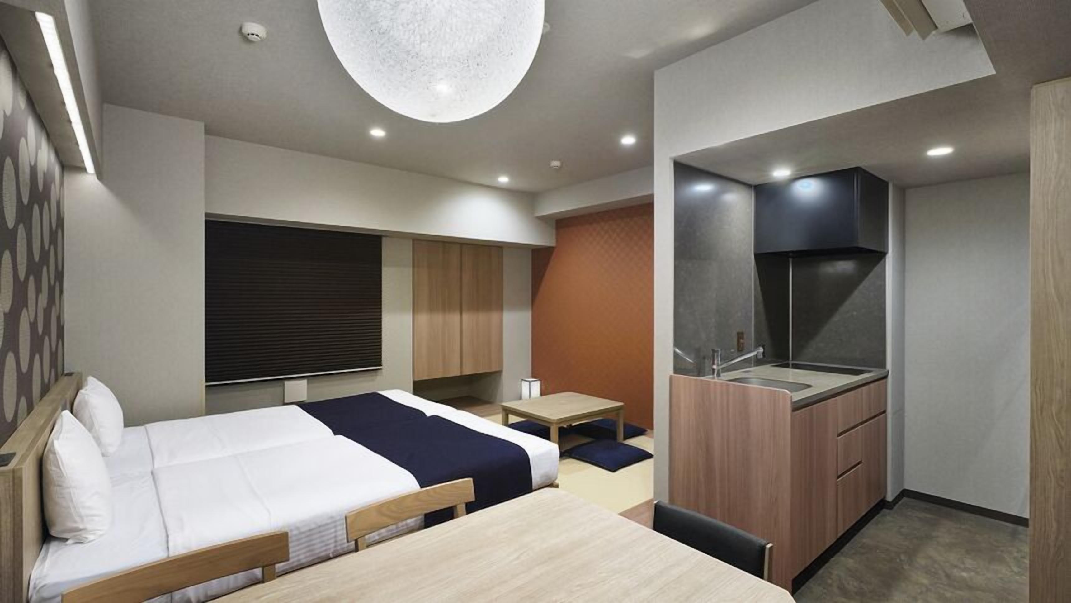 A modern hotel room with a bed, a desk, a kitchenette, and a large window.