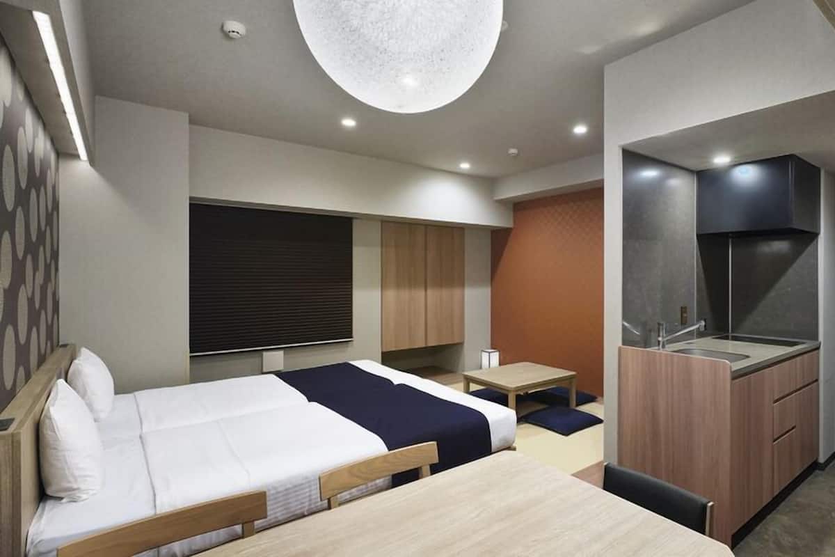 A modern hotel room with a bed, a desk, a kitchenette, and a large window.