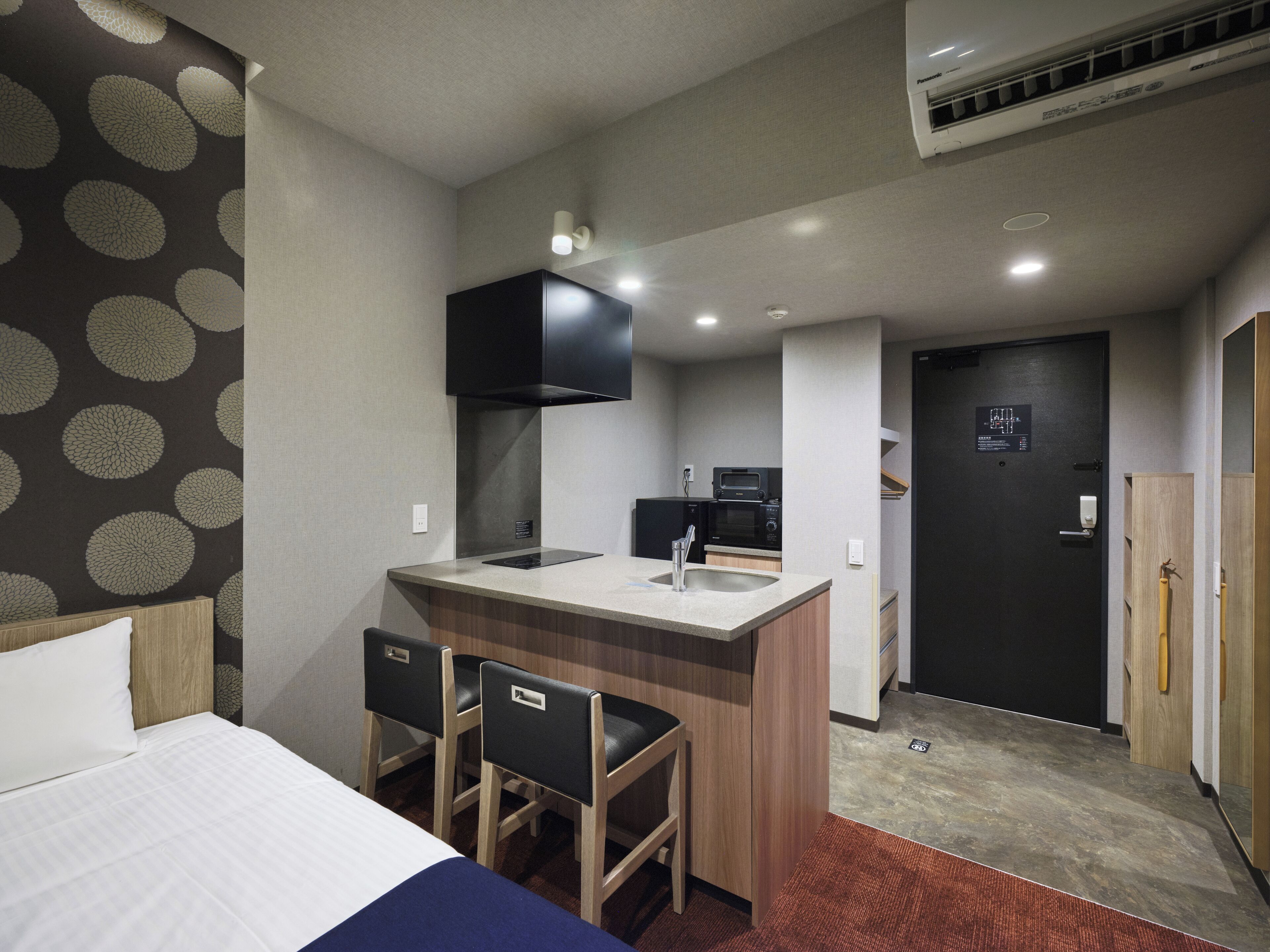 A compact hotel room with a bed, a small kitchenette, a bathroom, and a desk area.
