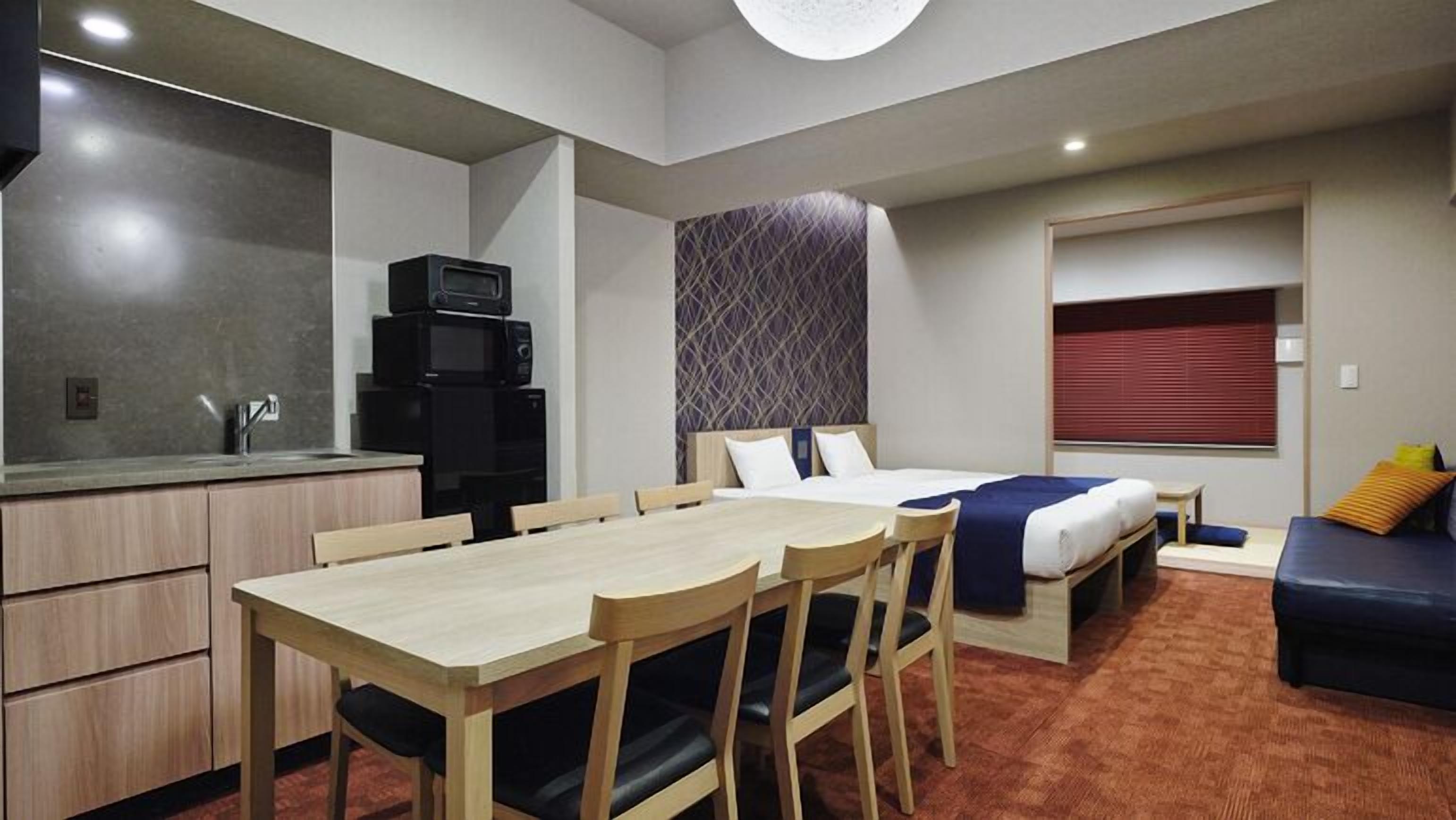 A compact hotel room with a kitchenette, a bed with a headboard, a sofa, and a window with blinds.