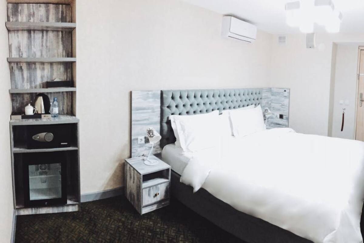 A hotel room with a large bed, a bedside table with a lamp, a wardrobe, a small desk with a chair, and a TV mounted on the wall.