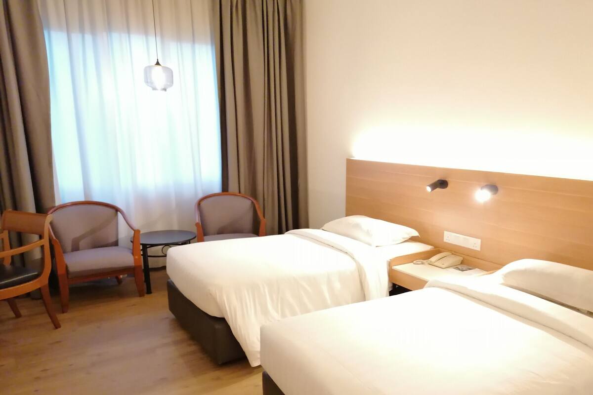 A hotel room with two beds, a small table, two chairs, a lamp, and a window with curtains.