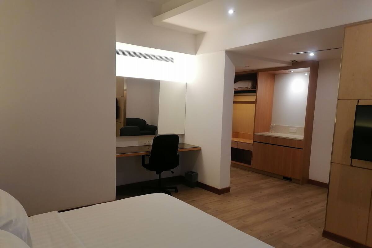 A compact hotel room with a bed, a desk, a chair, a kitchenette, and a wardrobe.