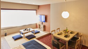 Ein Hotelzimmer mit einem Bett, einem Esstisch mit Stühlen, einem Fernseher und einem kleinen Tisch mit einer Vase und einer Schale.