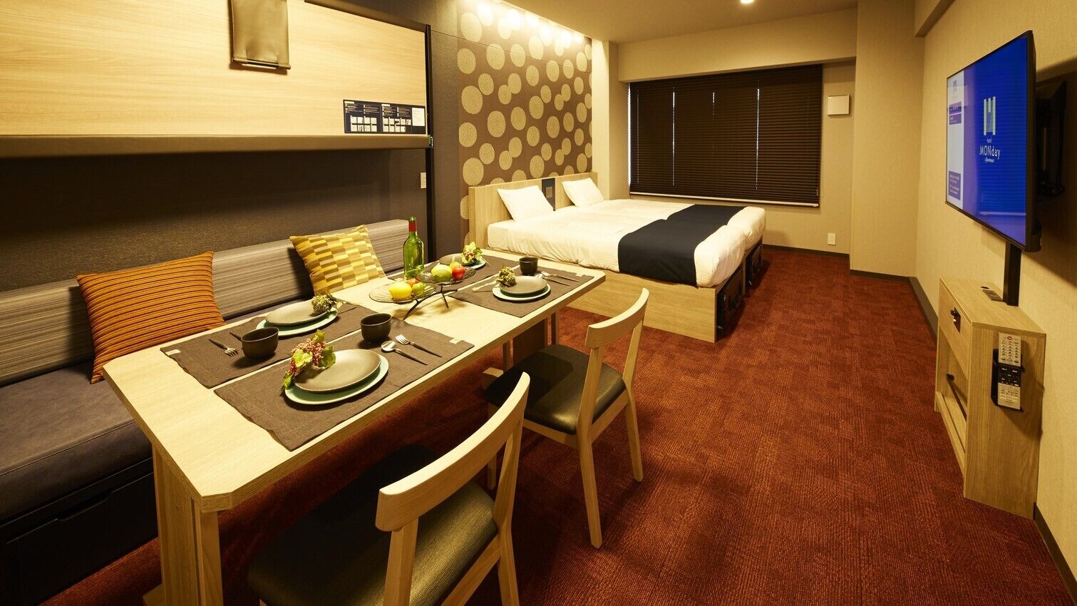 A compact hotel room with a dining table, two chairs, a sofa, a bed, a TV, and a wall-mounted control panel.