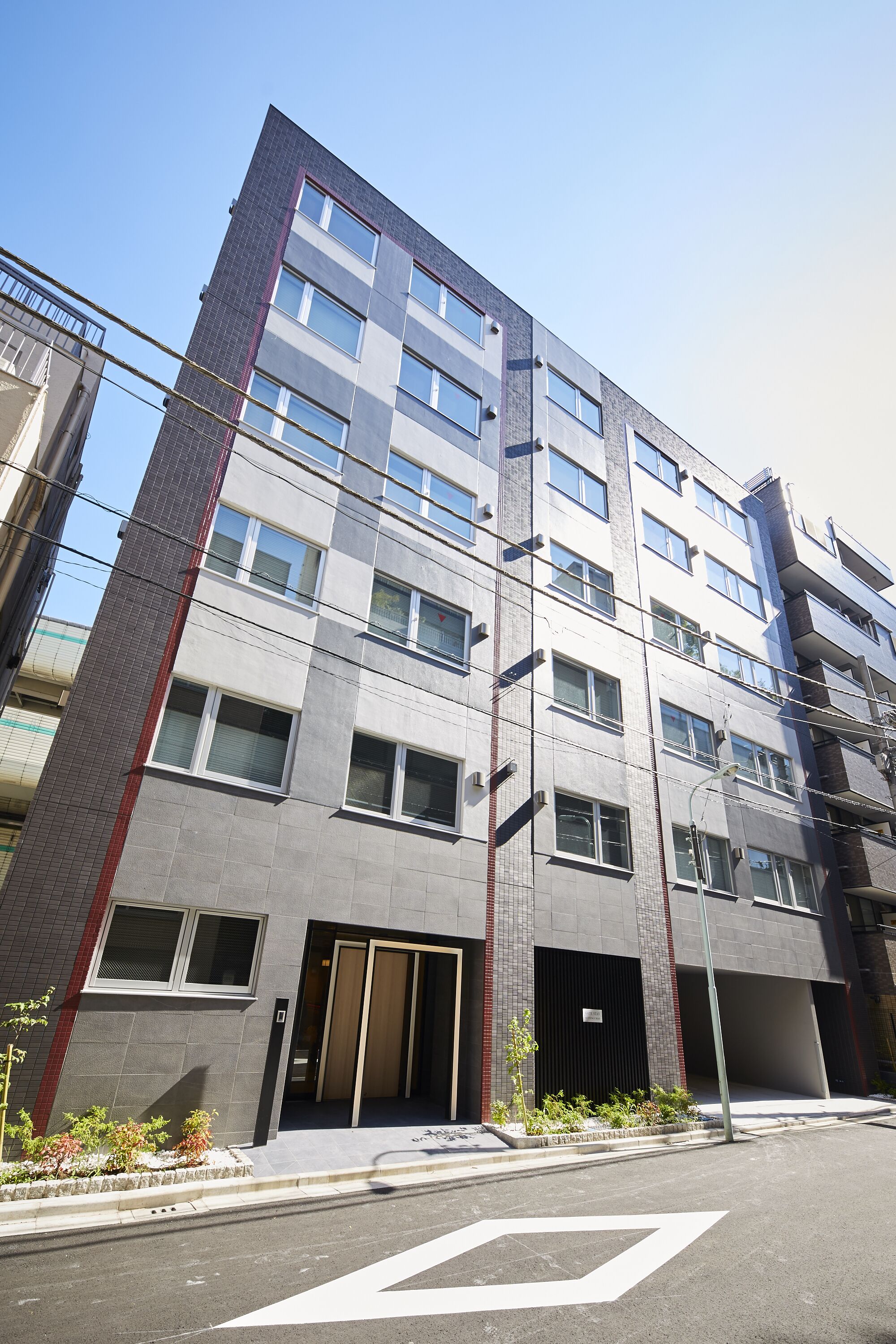 A multi-story building with a modern architectural design, featuring a mix of glass and brick facade, large windows, and a ground-level entrance with a small garden area.