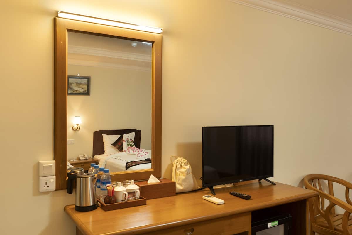 Quarto de hotel com uma secretária de madeira, televisão de ecrã plano, espelho, cama com cabeceira branca e uma pequena mesa de cabeceira com candeeiro e telefone.