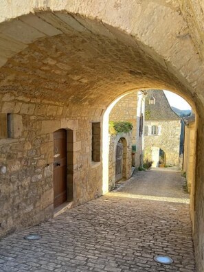 A narrow cobblestone street with stone buildings on either side, an archway, and a visible door on the left.