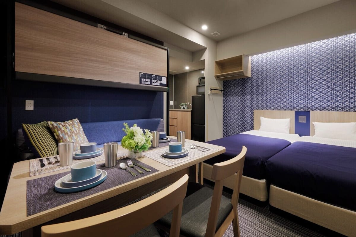 A compact hotel room with a kitchenette, a dining area, and a bed with a patterned headboard.