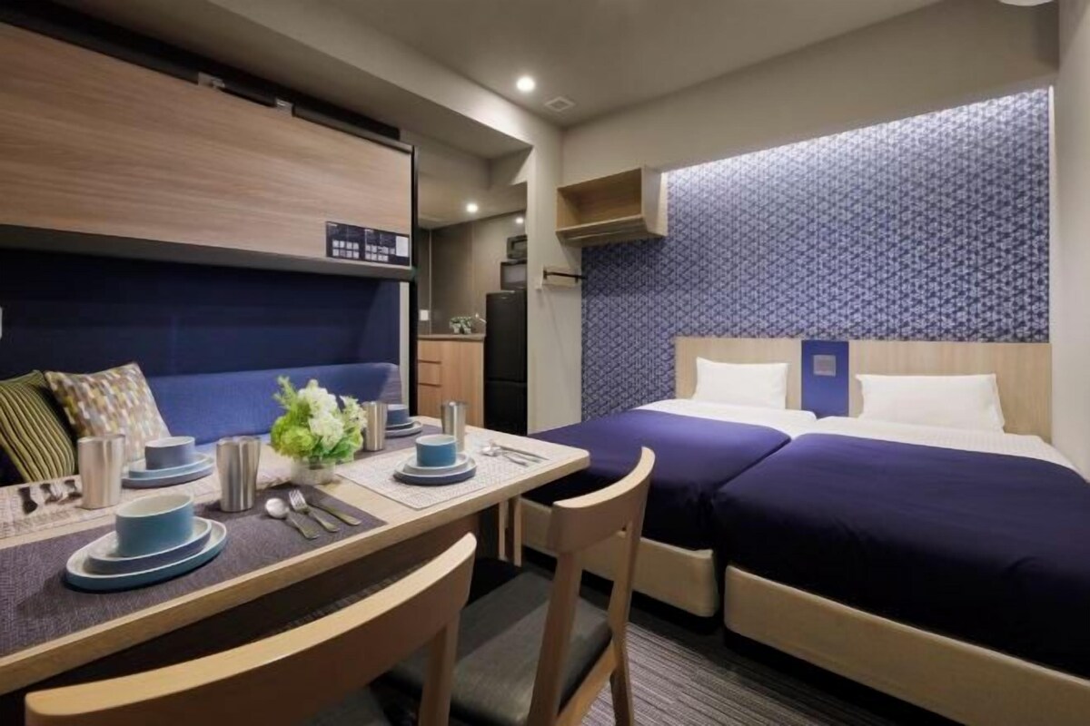 A hotel room with a bed, a desk with a chair, a small table with a vase of flowers, and a wall-mounted shelf.