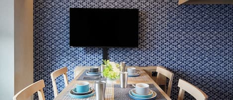 Un comedor con una mesa preparada para cuatro, un televisor de pantalla plana montado en la pared y papel tapiz estampado.