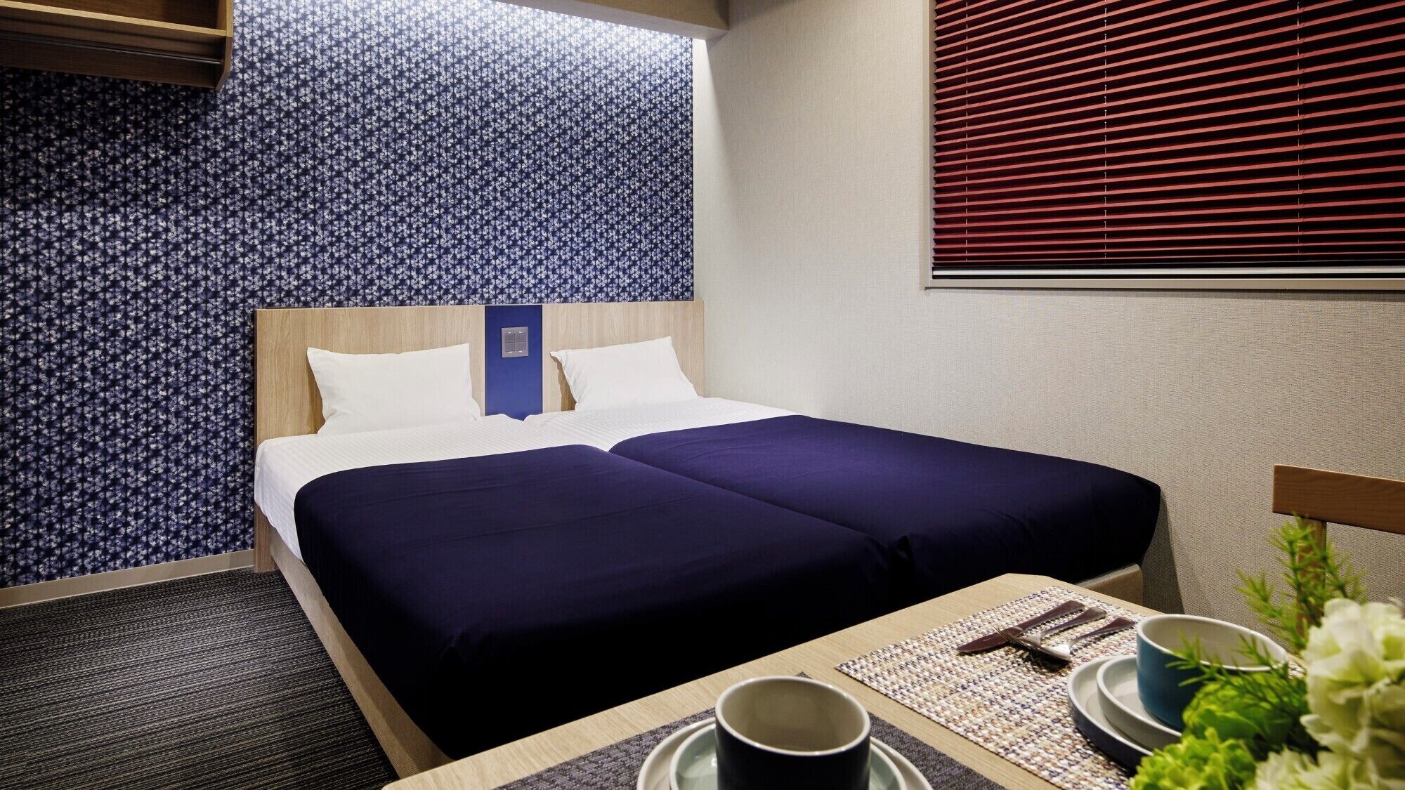Une chambre d’hôtel avec un lit, une table de chevet sur laquelle se trouvent une tasse et un bol, et une fenêtre avec des stores.