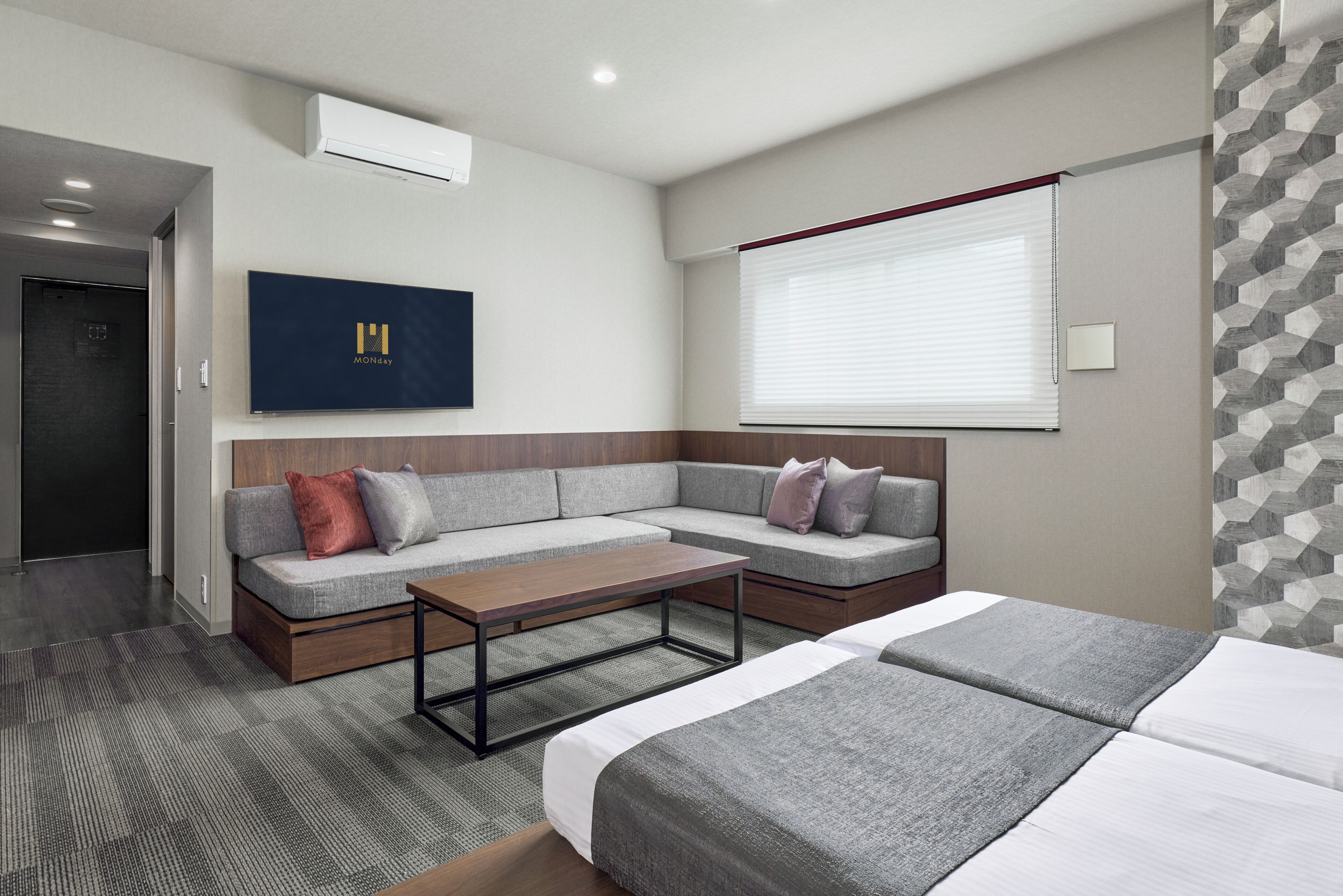 A modern hotel room with a sectional sofa, a bed, a coffee table, and a large window with blinds.