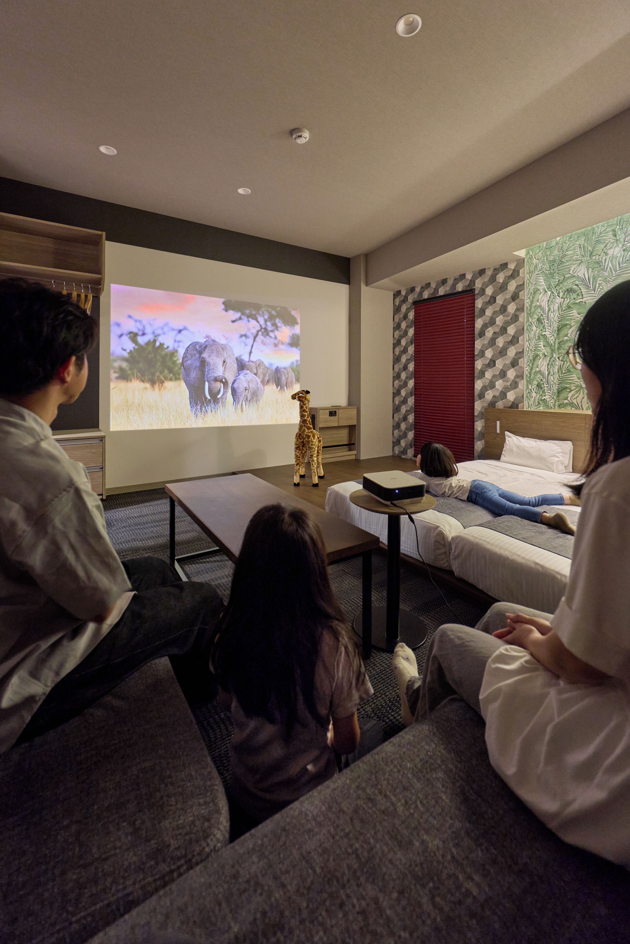 A living room with a large screen displaying an animal projection, a sofa, a coffee table, and a bed with a person lying down.